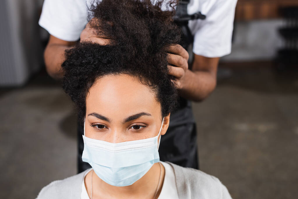 Joven cliente afroamericano en máscara médica sentado cerca de estilista sobre fondo borroso  - Foto, imagen