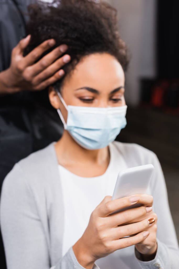Smartphone in hands of blurred african american client in medical mask near hairdresser  - Photo, Image