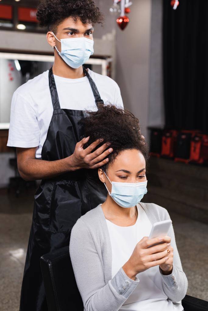 Femme utilisant un smartphone près de coiffeur afro-américain dans un masque médical  - Photo, image