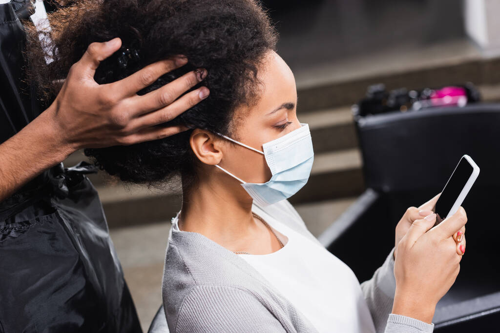 Afrikanisch-amerikanischer Kunde in medizinischer Maske mit Smartphone in der Nähe von Friseur in Schürze  - Foto, Bild