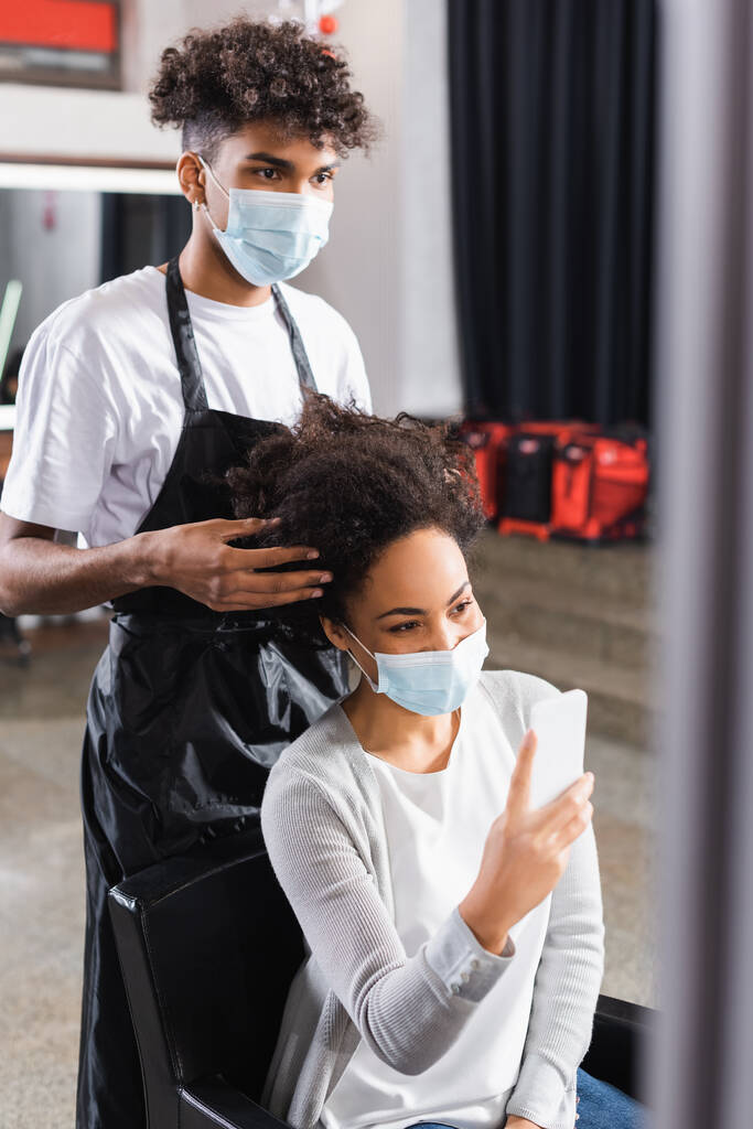 アフリカ系アメリカ人の美容師は、スマートフォンでクライアントの髪に触れる  - 写真・画像