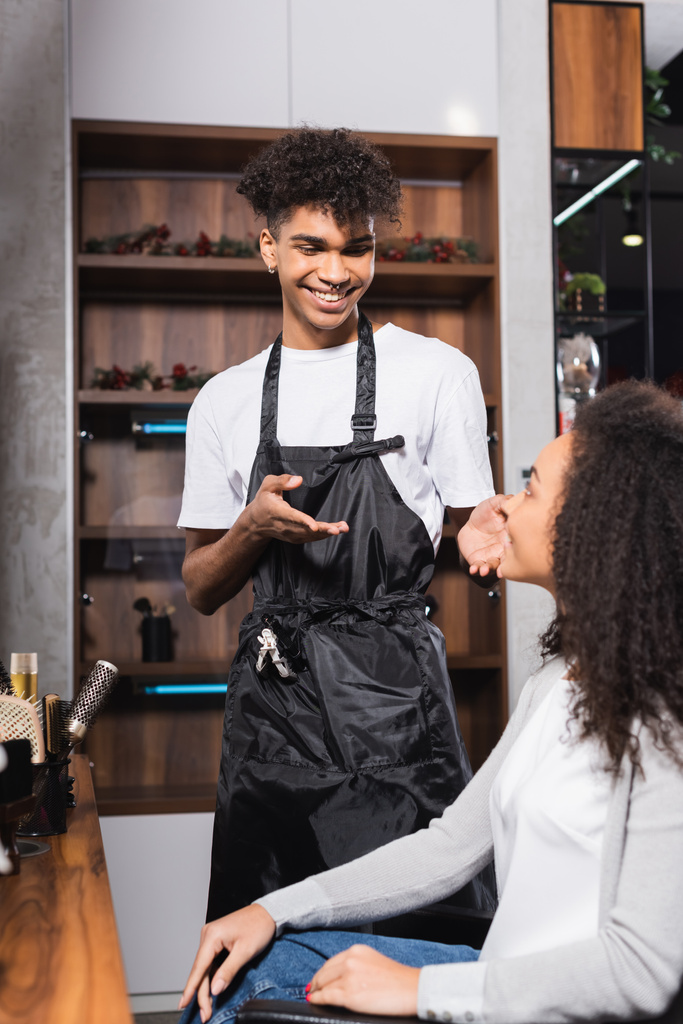 Afrykański amerykański fryzjer wskazujący uśmiechniętą kobietę na niewyraźnym pierwszym planie w salonie  - Zdjęcie, obraz
