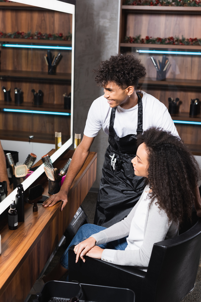 Позитивна афроамериканська перукарка, що дивиться на дзеркало біля клієнта в салоні.  - Фото, зображення