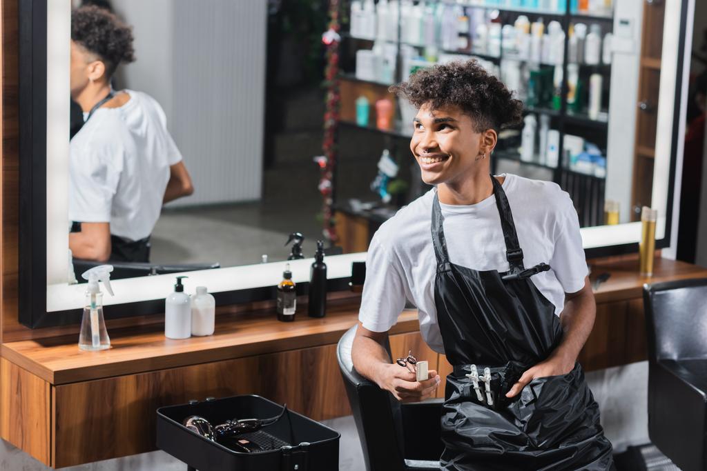 Sorrindo cabeleireiro afro-americano com tesoura e pente olhando para longe no salão  - Foto, Imagem
