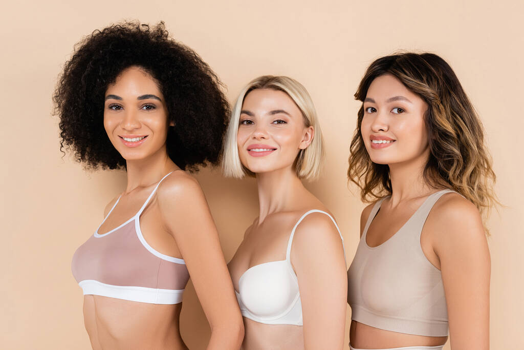 happy interracial women in bras smiling at camera on beige - Photo, Image