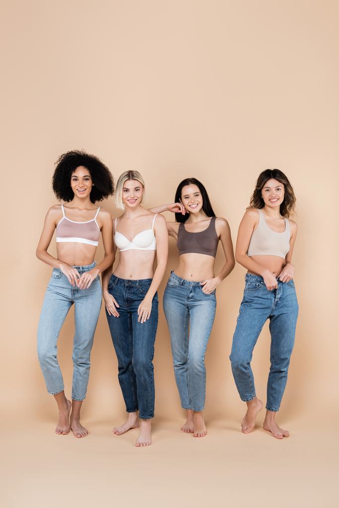 vista integrale di giovani donne multietniche in reggiseni e jeans in posa sul beige - Foto, immagini