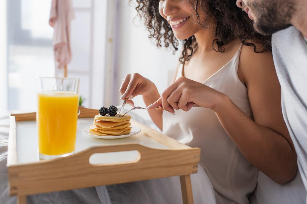 cropped view of happy african american woman holding cutlery near pancakes and boyfriend in bedroom  - Photo, Image