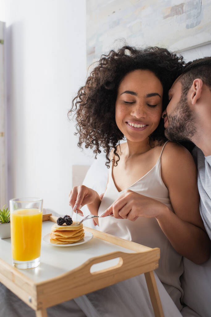 uomo barbuto baciare felice donna africana americana in possesso di posate vicino frittelle in camera da letto - Foto, immagini