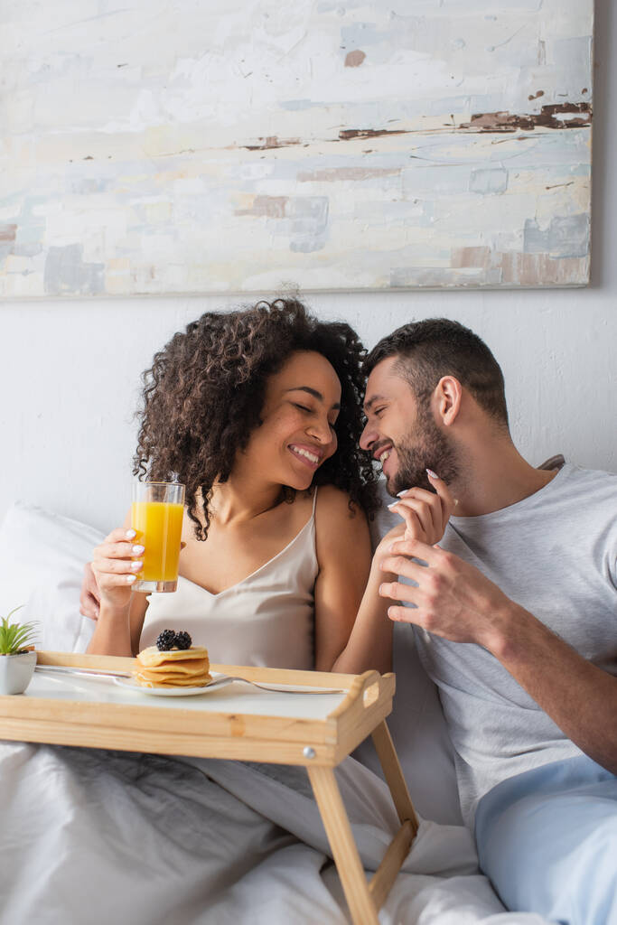 allegra donna afroamericana che tiene un bicchiere di succo d'arancia vicino a frittelle e fidanzato in camera da letto  - Foto, immagini