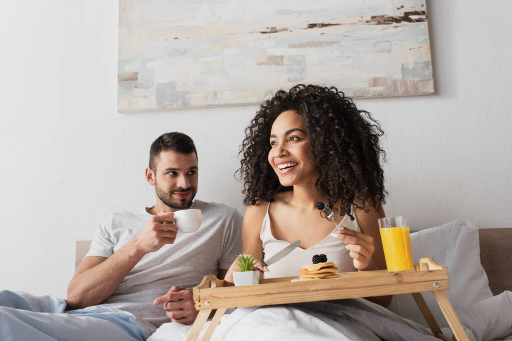 smiling african american woman holding fork with blackberry near pancakes on tray and boyfriend with cup of coffee - Photo, Image