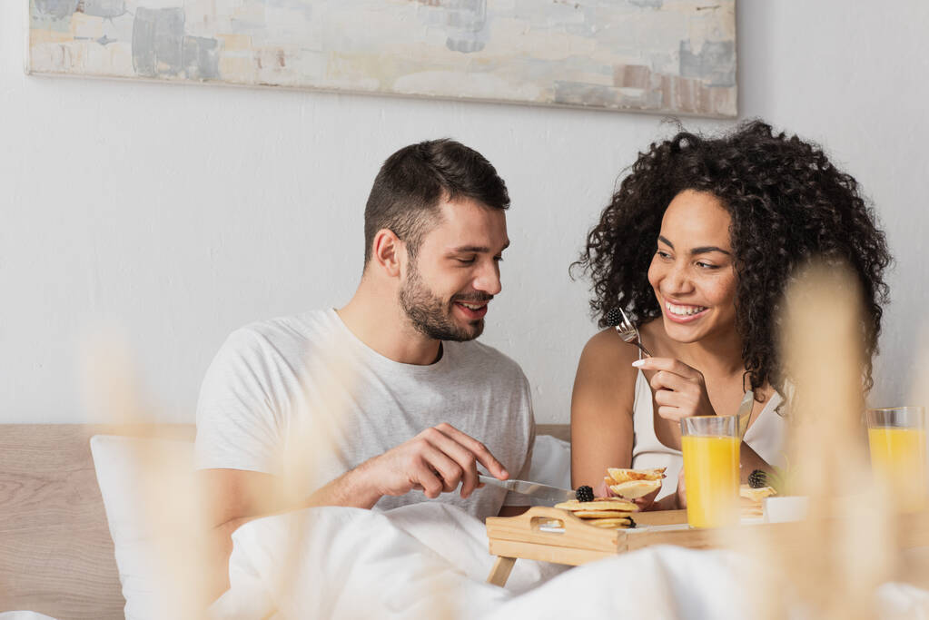 iloinen rotujenvälinen pari hymyilee ja ottaa aamiaisen makuuhuoneessa - Valokuva, kuva