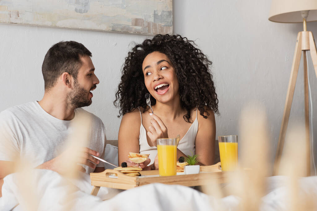 Sakallı adam Afrikalı Amerikalı kız arkadaşına bakıyor ve yatakta böğürtlen yiyor. - Fotoğraf, Görsel