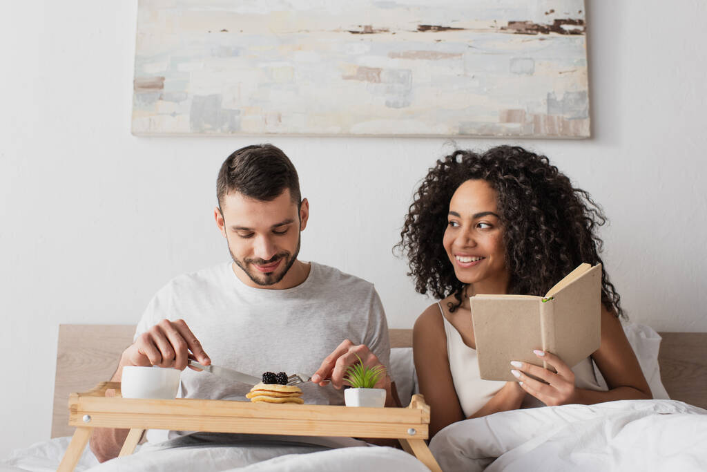 feliz Africano americano segurando livro perto namorado tomando café da manhã na cama - Foto, Imagem