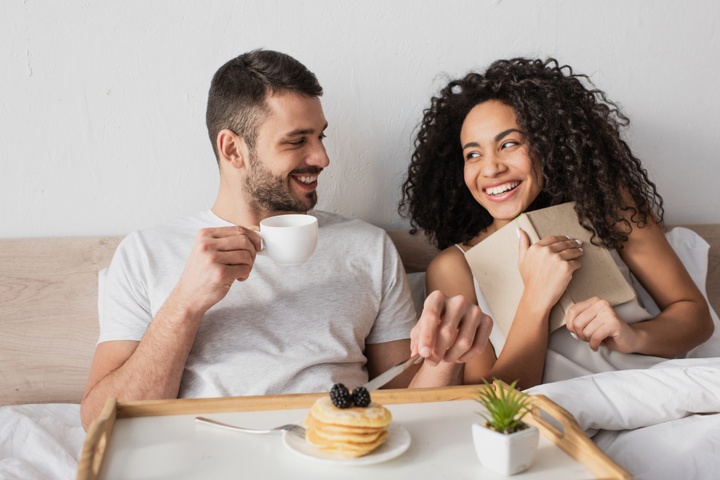 alegre Africano americano segurando livro perto feliz namorado tomando café da manhã na cama - Foto, Imagem