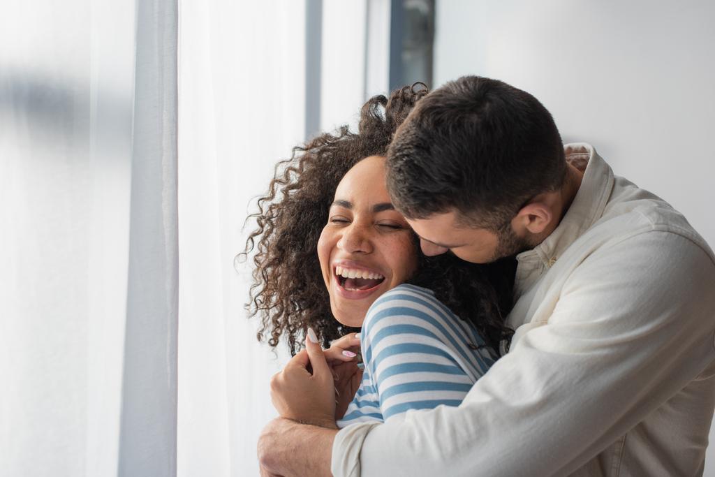 cheerful multiethnic couple hugging at home - Photo, Image