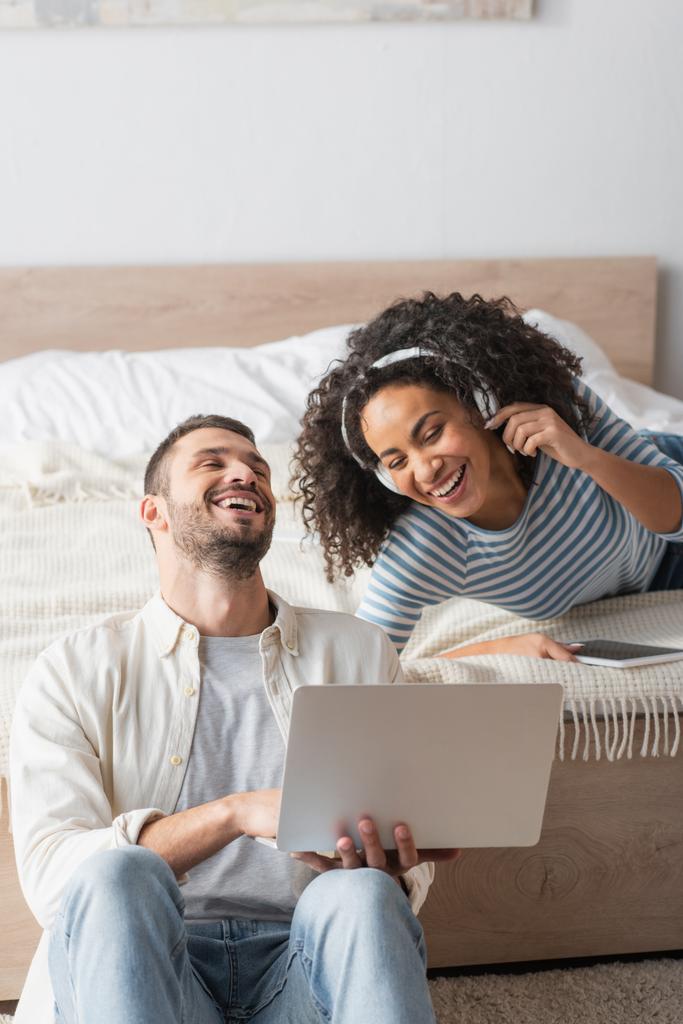pareja interracial riendo cerca de gadgets en el dormitorio - Foto, imagen