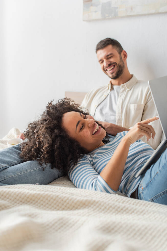 feliz afroamericana mujer acostada en la cama y señalando con el dedo a la computadora portátil cerca de novio  - Foto, imagen