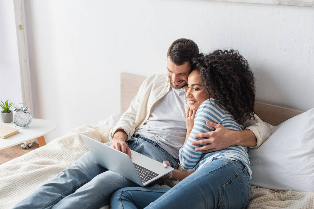 alegre interracial pareja chilling en cama y viendo comedia película en laptop en dormitorio - Foto, imagen