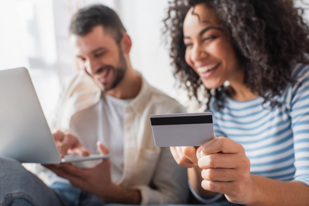 cartão de crédito na mão na mulher americana africana alegre perto do namorado com laptop no fundo borrado - Foto, Imagem