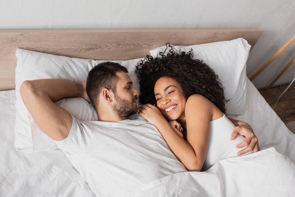 Yatakta gülümseyen Afrikalı Amerikalı kız arkadaşına bakan sakallı bir adamın yüksek açılı görüntüsü. - Fotoğraf, Görsel
