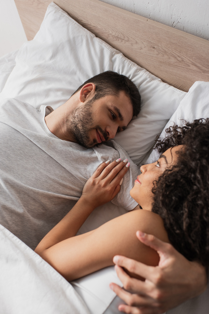 vue grand angle de couple interracial joyeux se regardant tout en étant couché dans le lit à la maison - Photo, image
