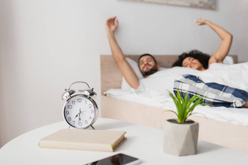 alarm clock, book, plant and smartphone on coffee table near interracial couple on blurred background  - Fotografie, Obrázek