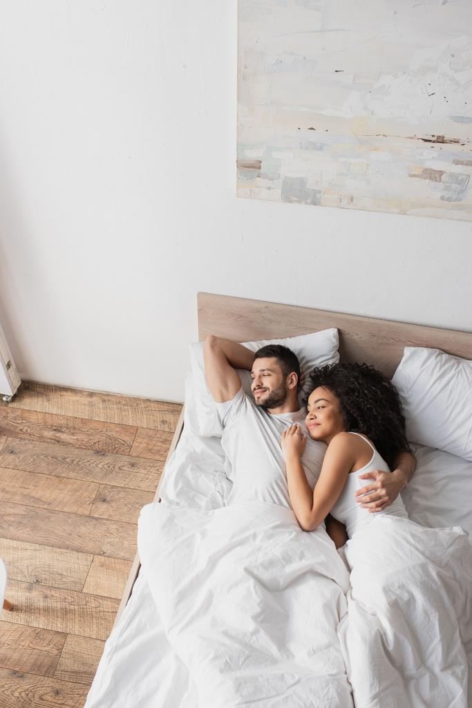 vista superior de positivo interracial casal abraçando enquanto deitado na cama - Foto, Imagem