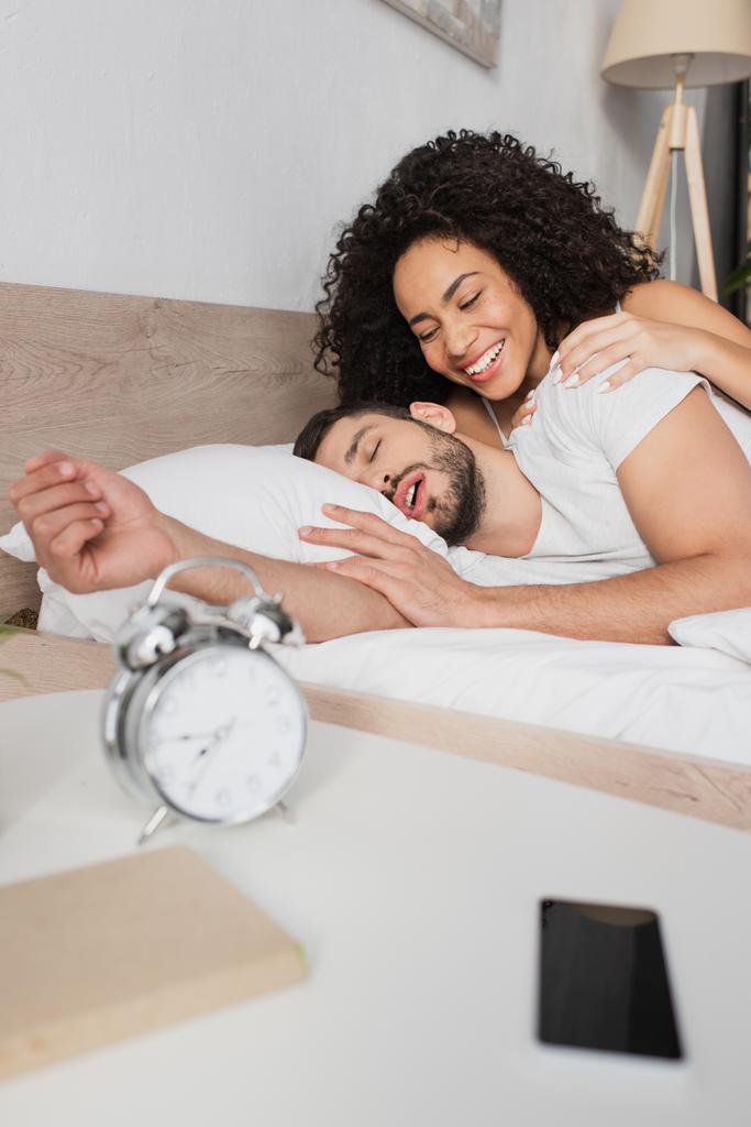 Mujer afroamericana sonriendo mientras mira al hombre barbudo durmiendo en la cama - Foto, imagen