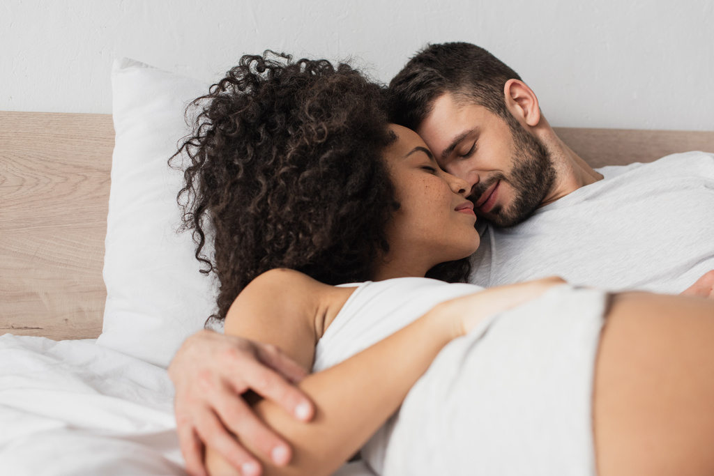 веселый межрасовый мужчина и женщина обнимаются, лежа в постели - Фото, изображение