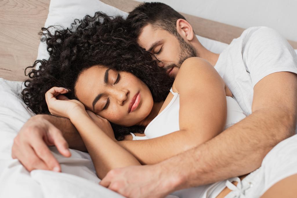 πολυεθνικός άντρας και γυναίκα αγκαλιάζονται ενώ κοιμούνται στο κρεβάτι - Φωτογραφία, εικόνα