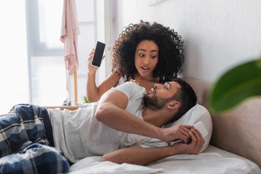 surprised african american woman looking at boyfriend while holding smartphone with blank screen in bedroom - Photo, Image