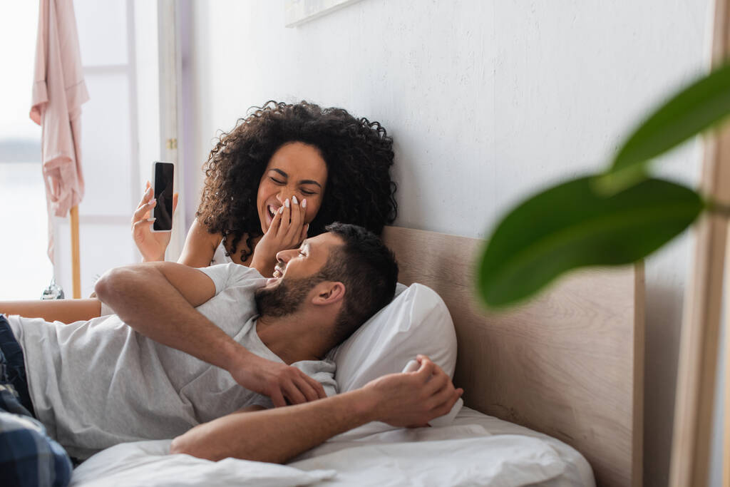 donna afroamericana che ride e guarda il fidanzato mentre tiene lo smartphone con schermo bianco in camera da letto con primo piano sfocato - Foto, immagini