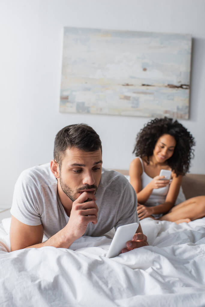 homme barbu couché sur le lit et textos sur smartphone près de femme afro-américaine sur fond flou  - Photo, image
