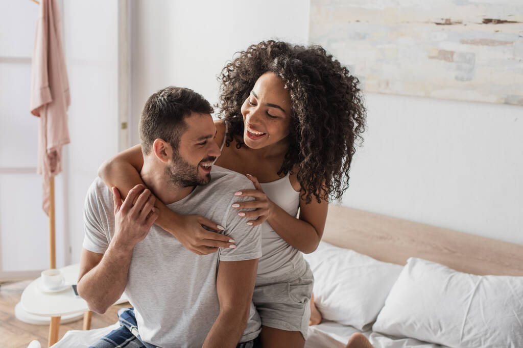curly african american woman hugging positive boyfriend in bedroom - Foto, afbeelding