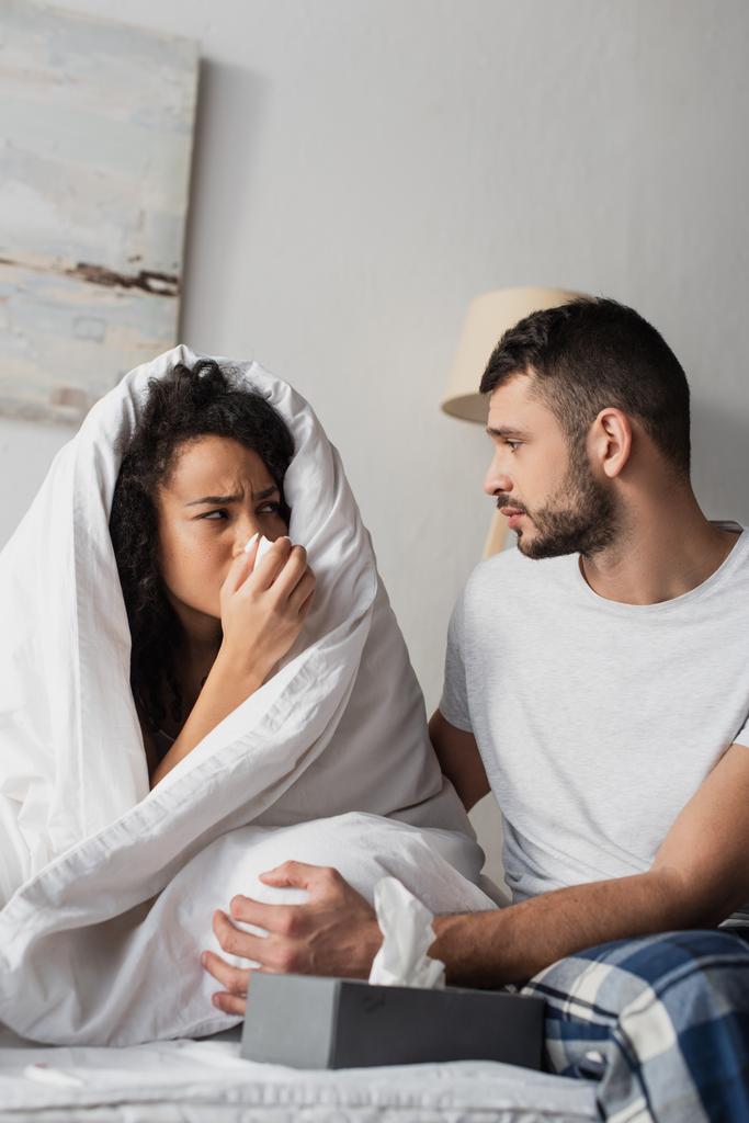 sick african american woman under blanket sneezing in napkin near worried boyfriend  - Fotografie, Obrázek