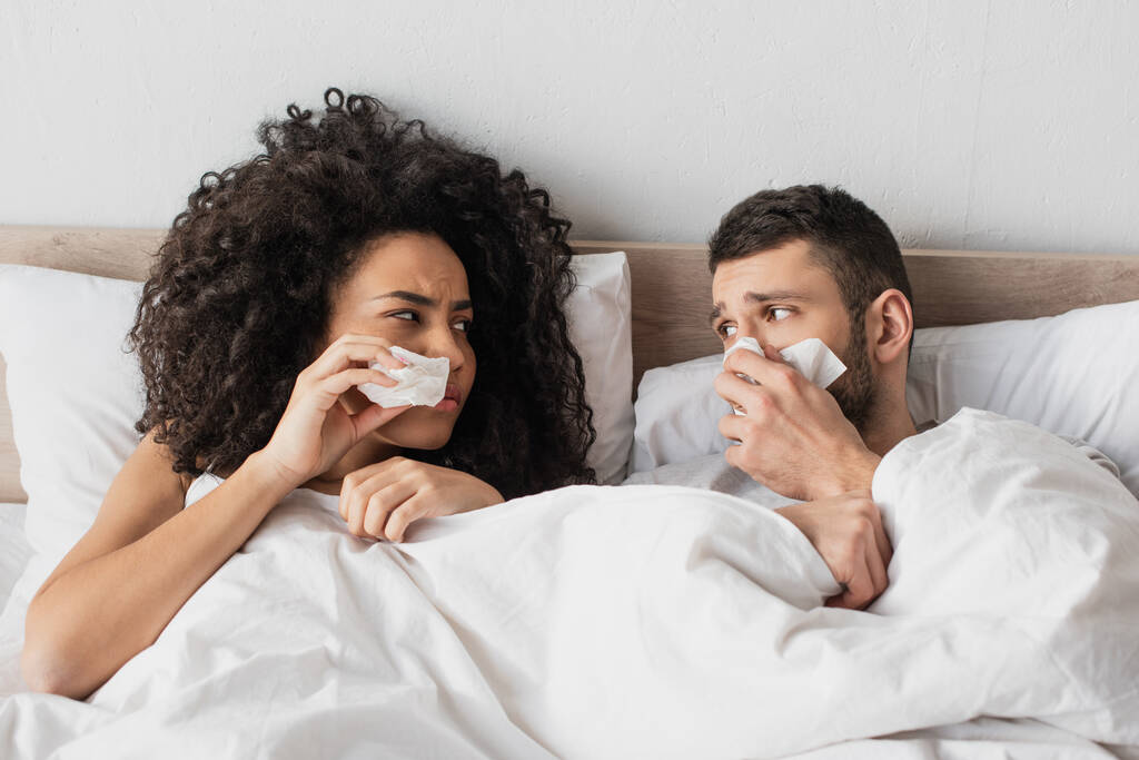 sick interracial couple holding napkins and sneezing while looking at each other - Photo, Image