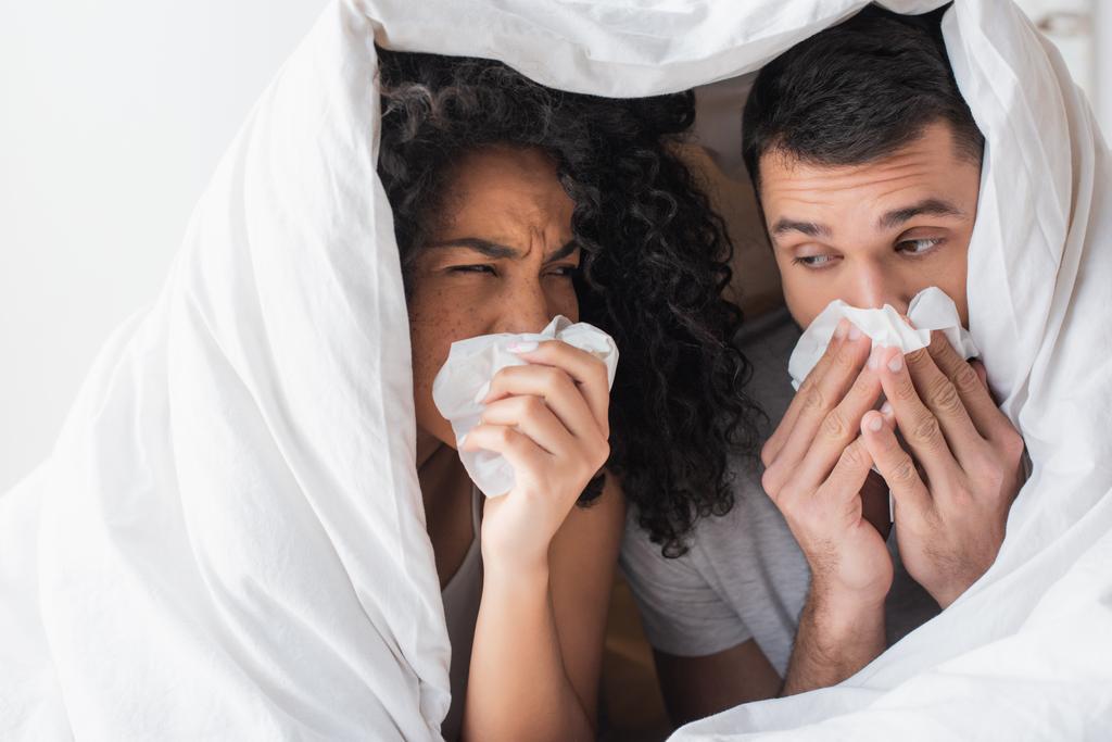 interracial sick couple under blanket sneezing in napkins  - Foto, afbeelding