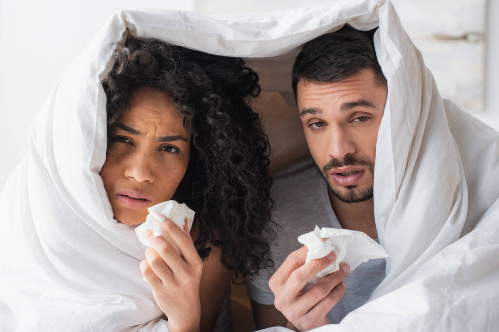 межрасовая больная пара под одеялом держит салфетки и смотрит в камеру - Фото, изображение