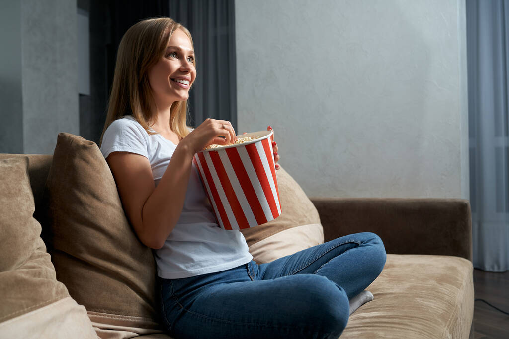 Uśmiechnięta kobieta ogląda film w telewizji i je popcorn - Zdjęcie, obraz