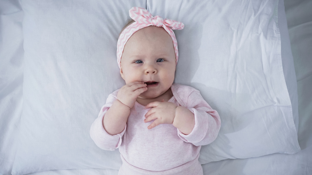 top view of baby girl in headband with bow lying on bed with white bedding - Photo, Image