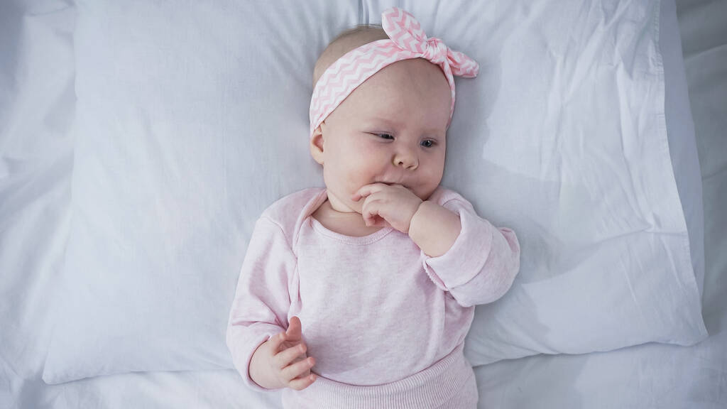 bovenaanzicht van baby met hoofdband liggend op bed en zuigvingers - Foto, afbeelding