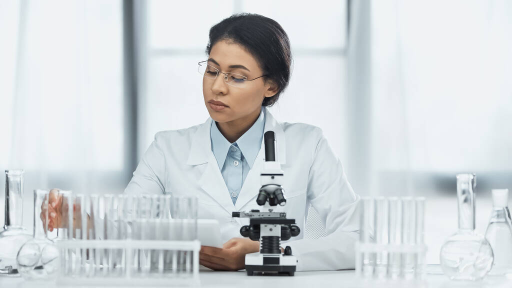 científico afroamericano en gafas tomando frasco cerca del microscopio en laboratorio  - Foto, Imagen