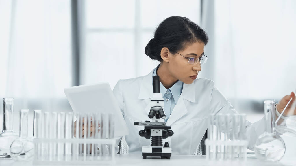 scientifique afro-américain dans des lunettes tenant tablette numérique près du microscope et des éprouvettes en laboratoire  - Photo, image