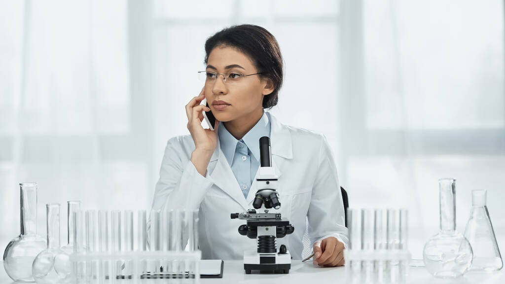 scienziato afroamericano in occhiali che parla su smartphone vicino al microscopio in laboratorio  - Foto, immagini