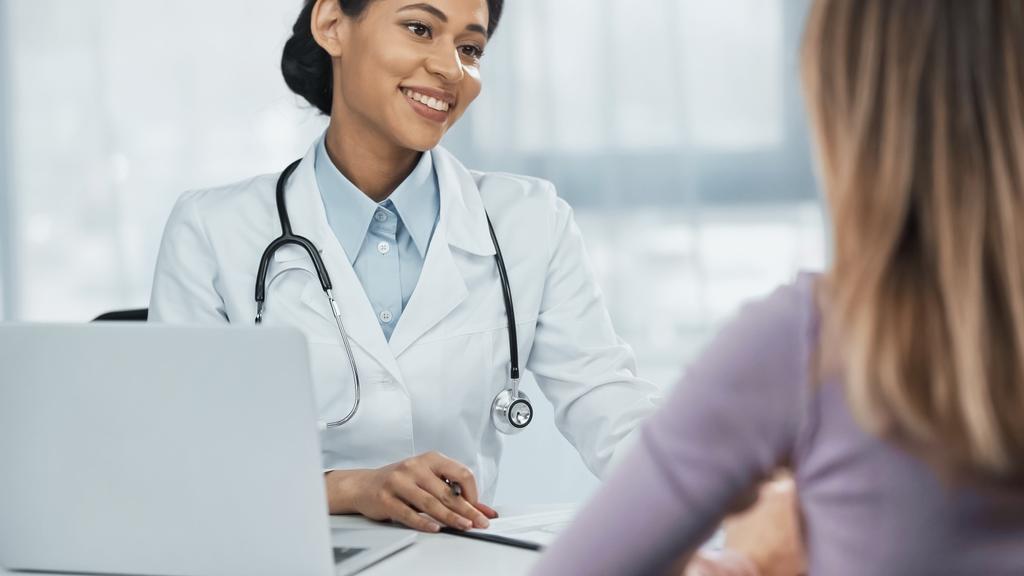 médico afroamericano feliz en bata blanca hablando con paciente borroso cerca del ordenador portátil en el escritorio - Foto, Imagen
