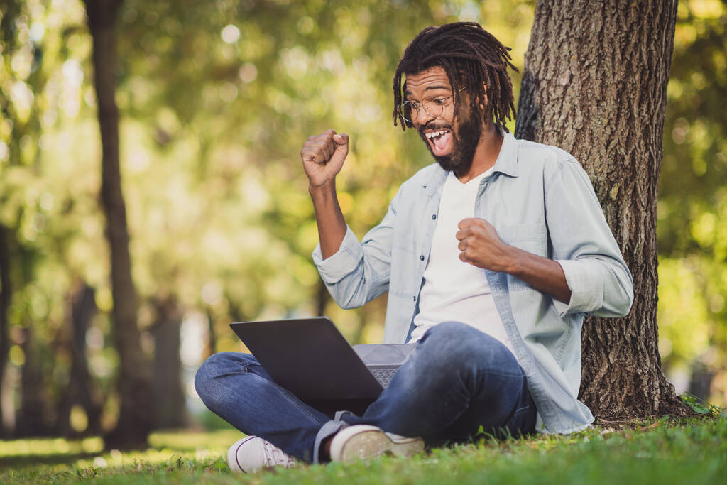 pleine taille photo de afro américain attrayant fou assis dans le parc herbe arbre lever poings gagner ordinateur à l'extérieur en plein air - Photo, image