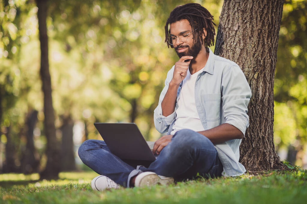 Retrato de bonito alegre focado cara viajante blogueiro sentado na grama verde usando laptop trabalhando dia ensolarado fora ao ar livre - Foto, Imagem
