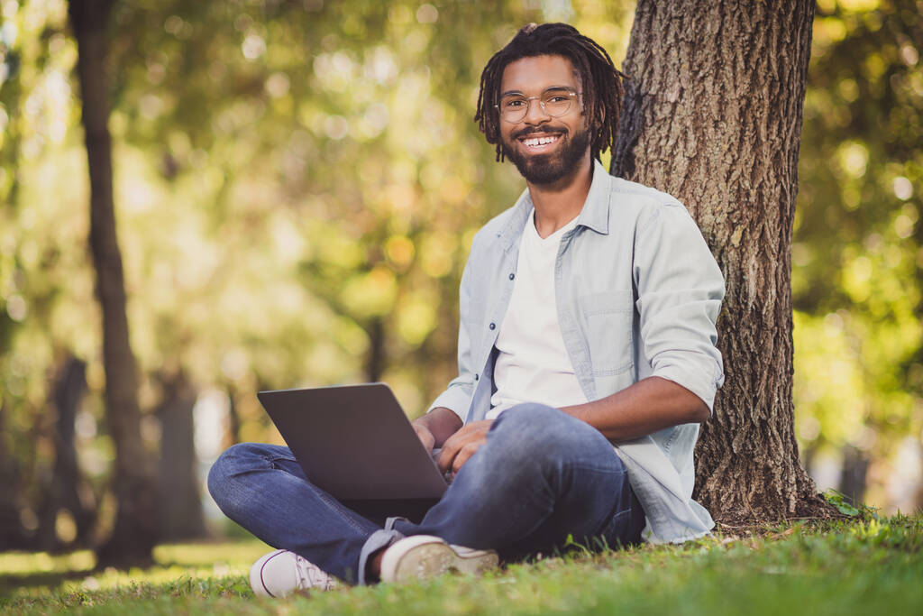 Full length φωτογραφία του γοητευτικό σκούρο δέρμα ευτυχισμένος άνθρωπος κάθονται δέντρο στο laptop έργο πάρκο καλή διάθεση έξω σε εξωτερικούς χώρους - Φωτογραφία, εικόνα