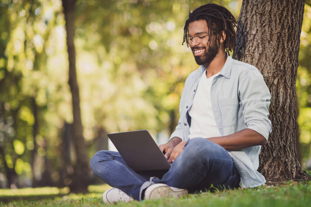 Fotoporträt eines Mannes mit Brille lächelnd im grünen Park sitzend und lächelnd am Laptop arbeitend - Foto, Bild