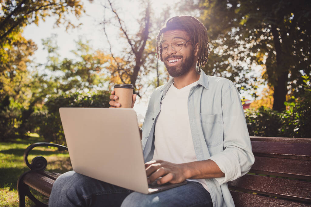 Kahverengi saçlı mutlu adam kahve bardağı tutuyor park bankında oturuyor bilgisayar dışarıda gözlük takıyor. - Fotoğraf, Görsel